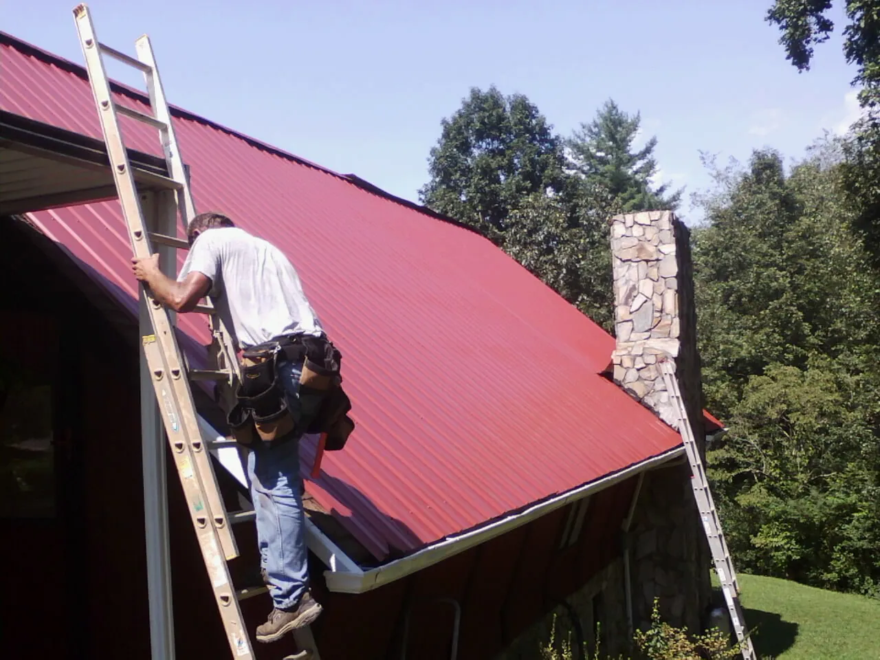 Roofer on a ladder beside a red metal roof and stone chimney — Soffit & Fascia Repair by Roofing Brothers in Daufuskie Island